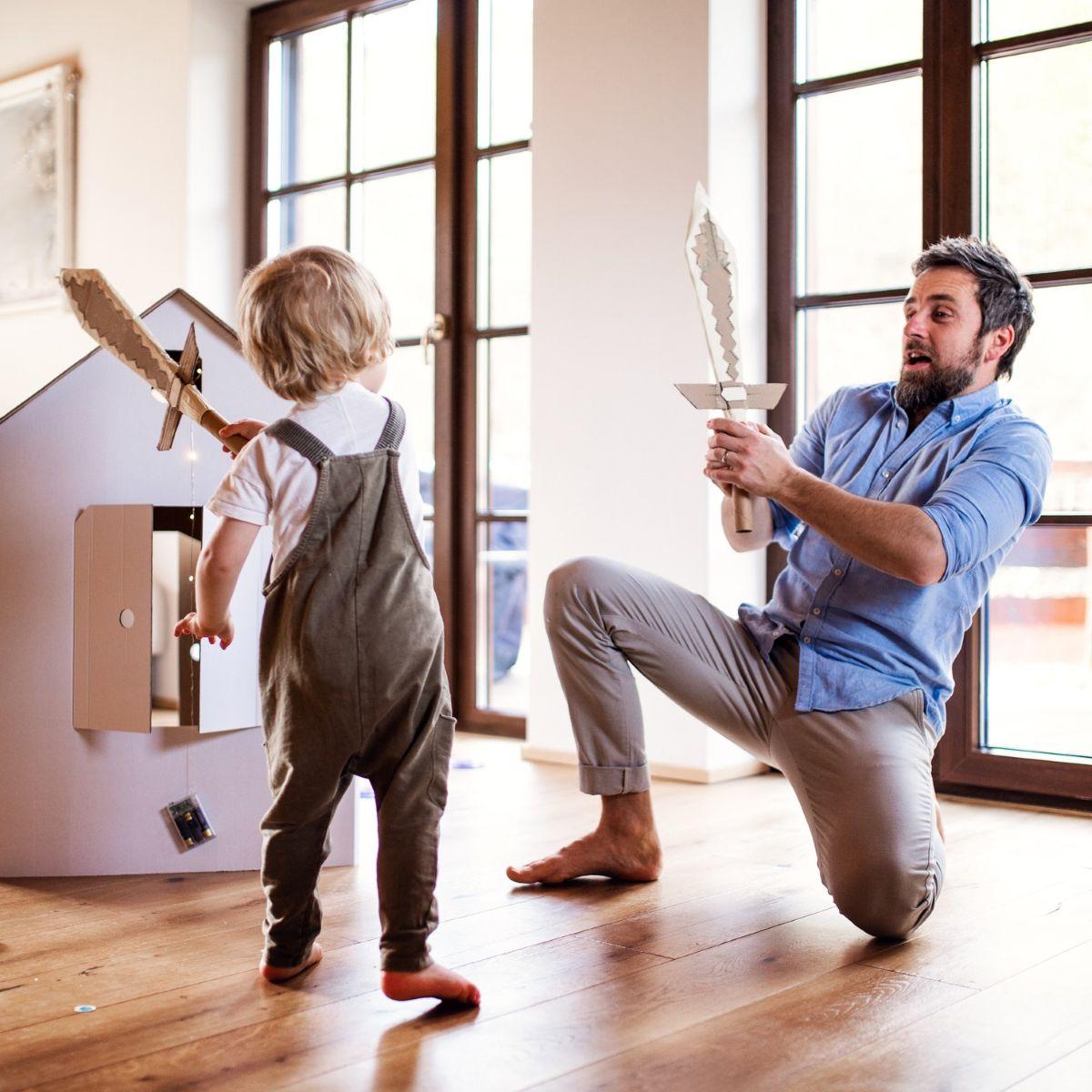 A toddler boy and father with carton swords playing indoors at home.