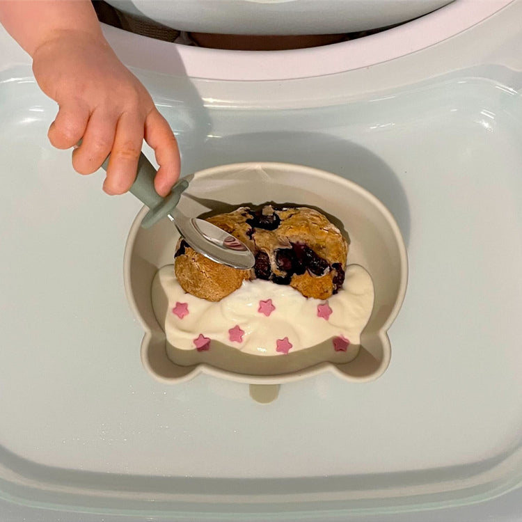 Baby eating solid food in bowl with spoon by herself