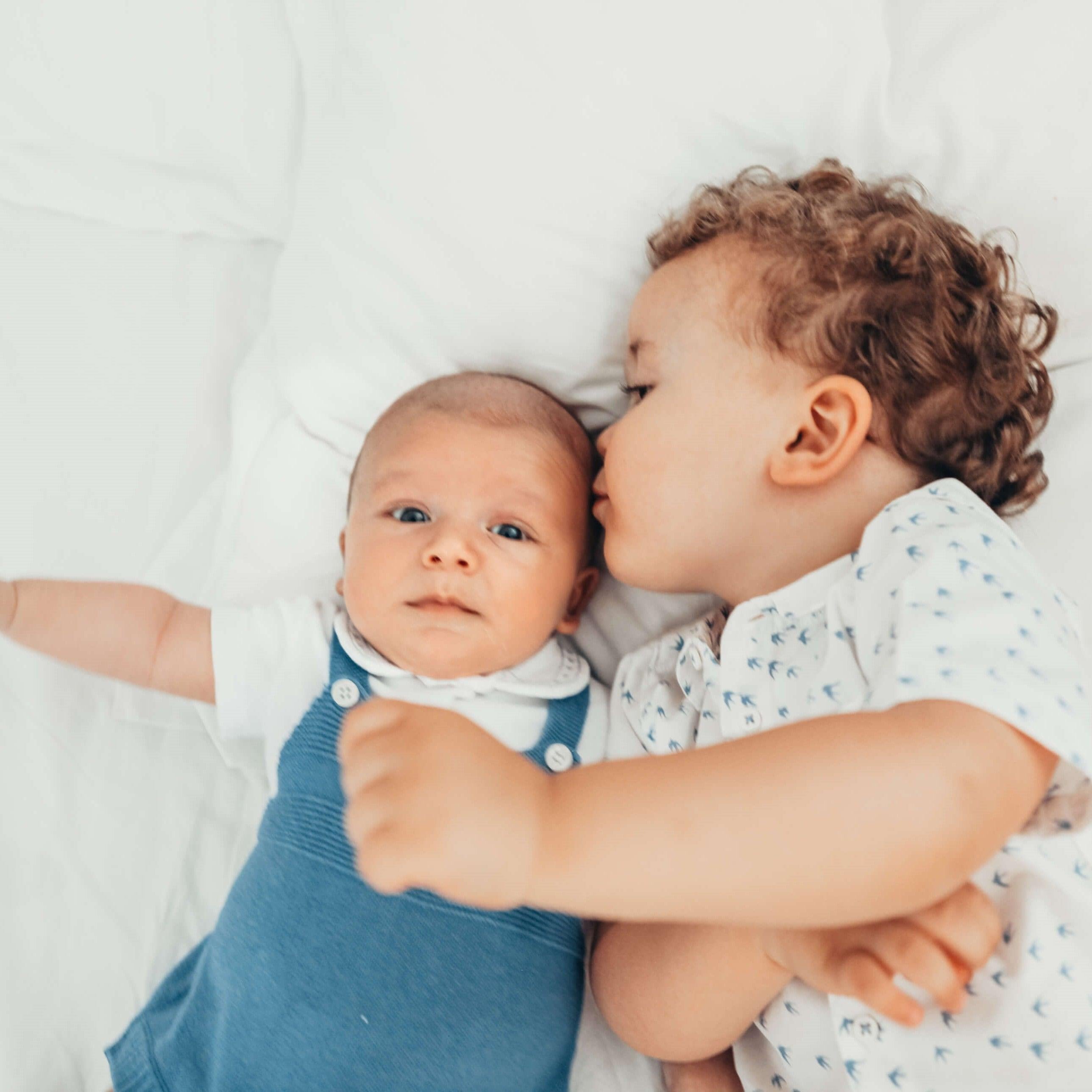 A baby is kissing her sibling on the bed.