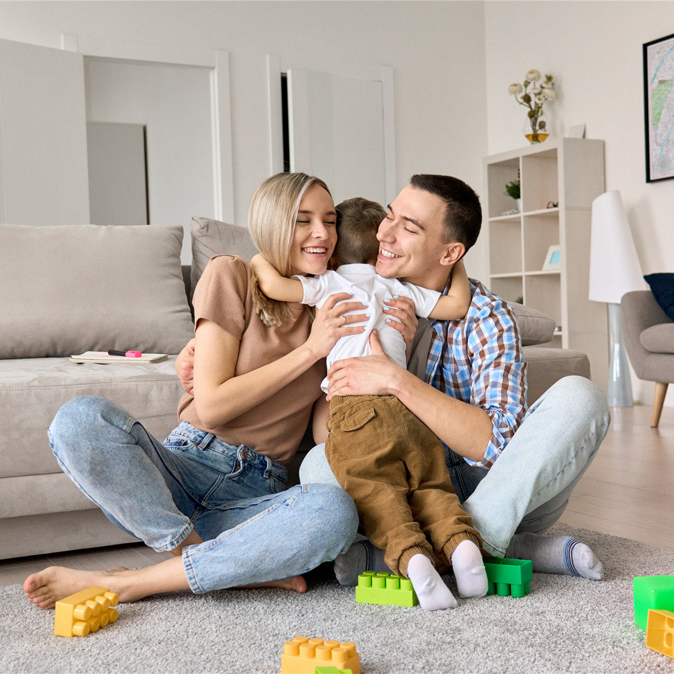 Happy young family couple having fun hugging cute small kid boy sitting on floor in modern living room